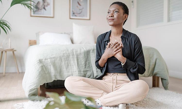 Woman with hands on her chest sitting on the floor in her bedroom in meditation.